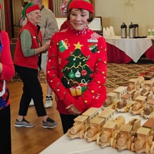 A senior resident dressed in Christmas attire is standing with purpose by a table of wooden trains and cars with other residents in the background.